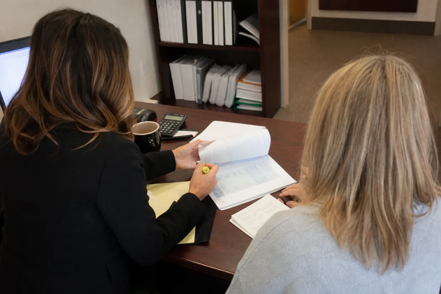 Agent in a black blazer flips through a policy statement with a highlighter beside a blonde client at a wood desk, with legal pad notes and a binder bookshelf behind